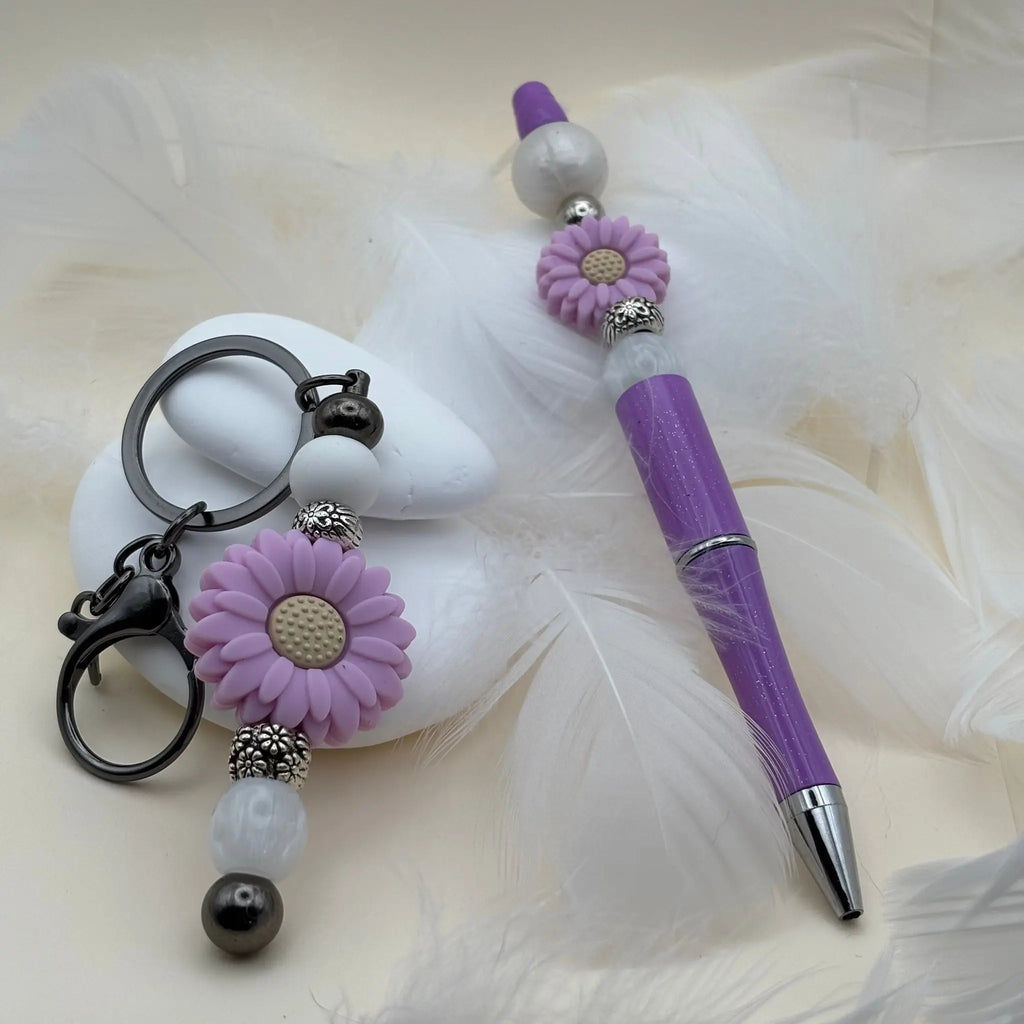 Stylo violet décoré de deux fleurs mauves et perles blanches, accompagné d’un porte-clés assorti avec une grande fleur mauve