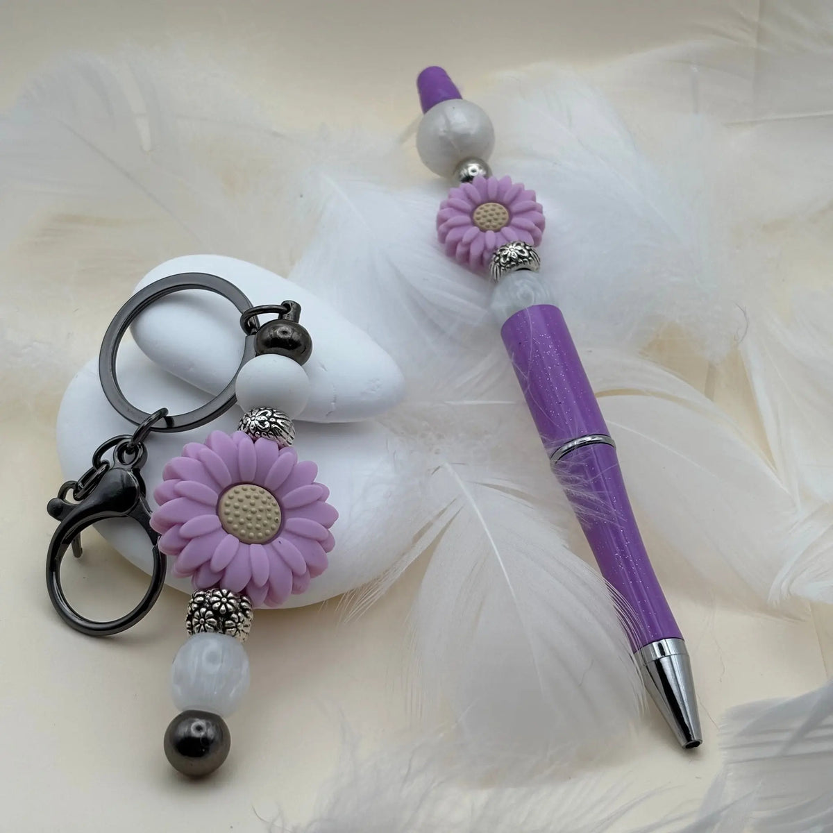 Stylo violet décoré de deux fleurs mauves et perles blanches, accompagné d’un porte-clés assorti avec une grande fleur mauve