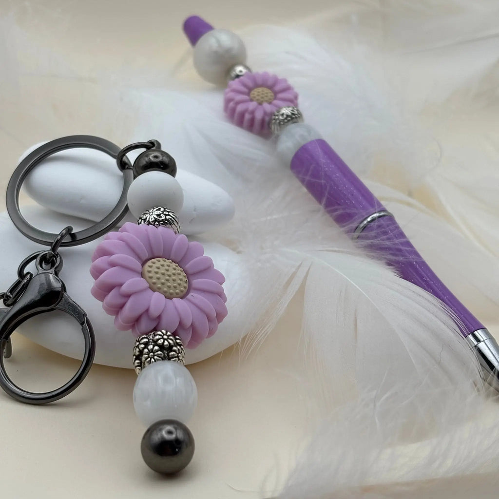 Stylo violet et blanc décoré de fleurs mauves, accompagné d’un porte-clés assorti posé sur des plumes blanches.