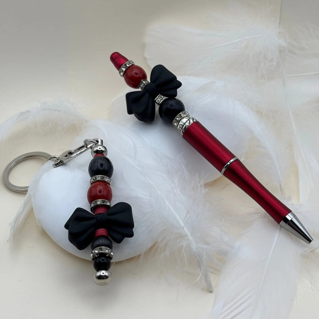 Stylo rouge décoré de perles noires et rouges avec nœuds noirs, accompagné d’un porte-clés assorti sur fond de plumes blanches