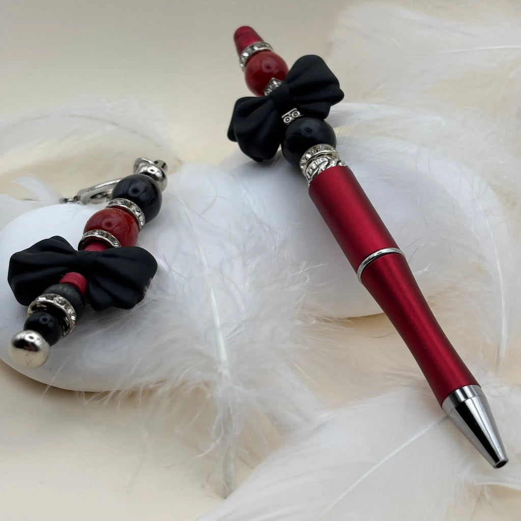 Stylo rouge décoré de nœuds noirs et perles noires et rouges, avec un porte-clés assorti sur fond de plumes blanches