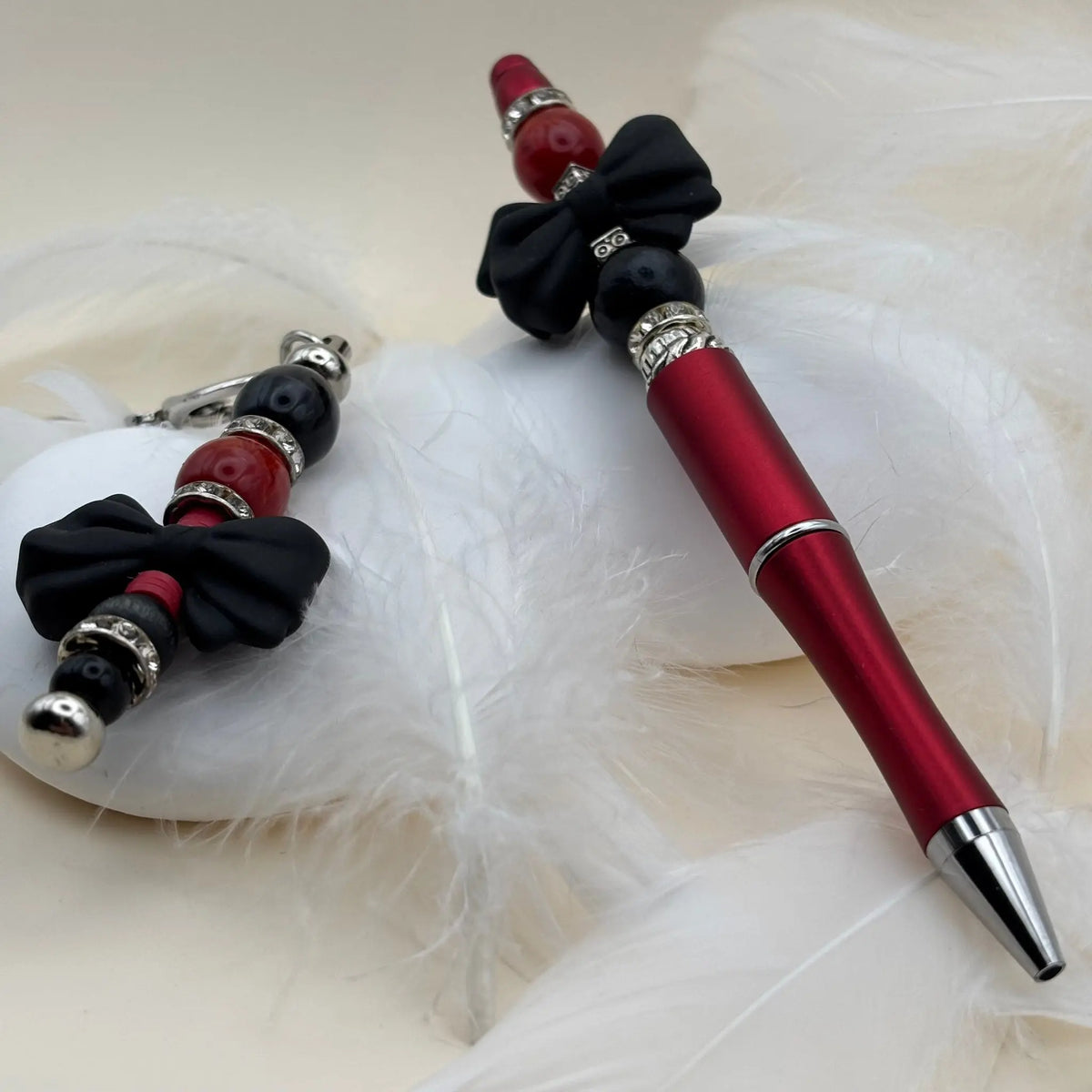 Stylo rouge décoré de nœuds noirs et perles noires et rouges, avec un porte-clés assorti sur fond de plumes blanches