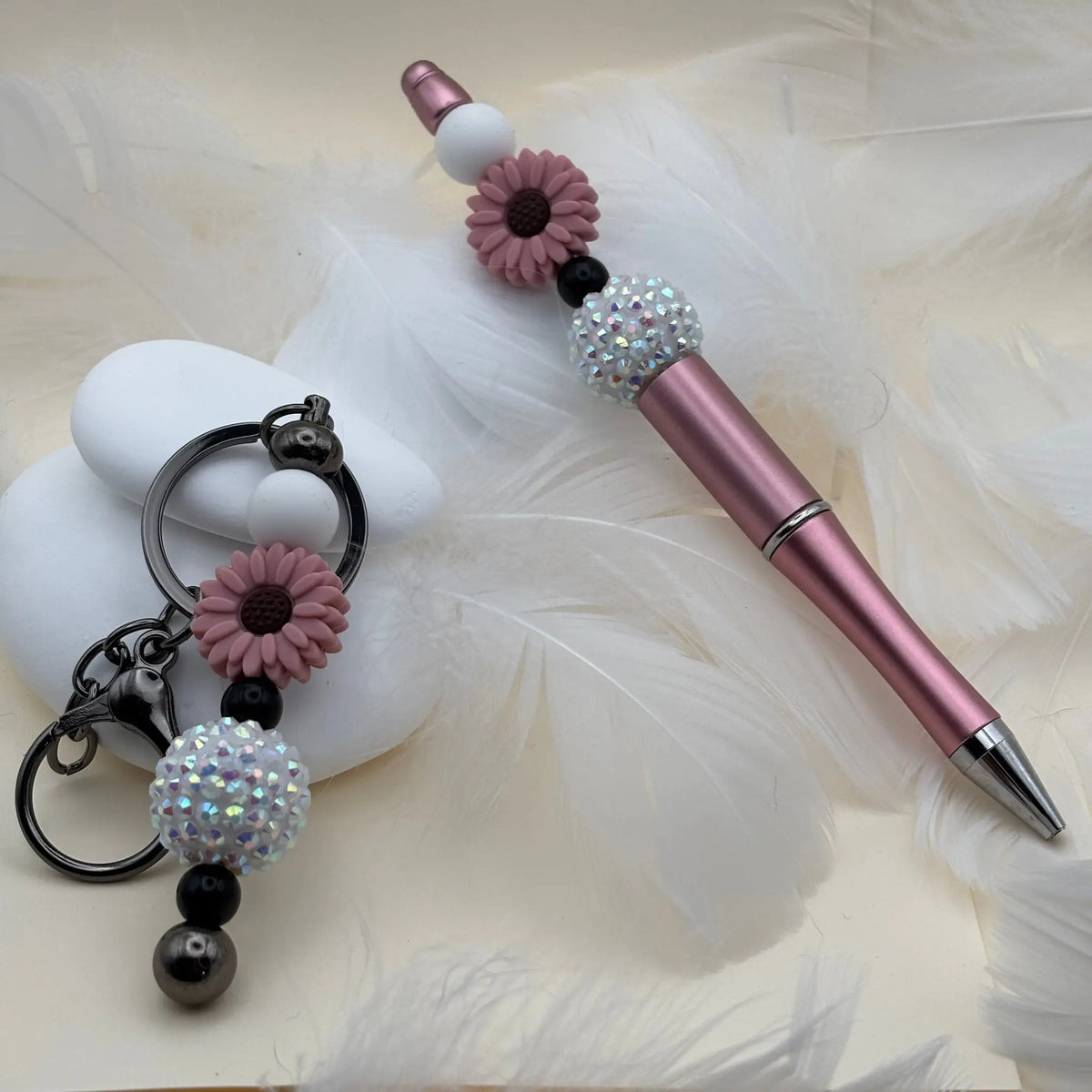 Stylo rose brillant décoré de perles blanches, noires et fleurs mauves, accompagné d’un porte-clés assorti posé sur des plumes blanches