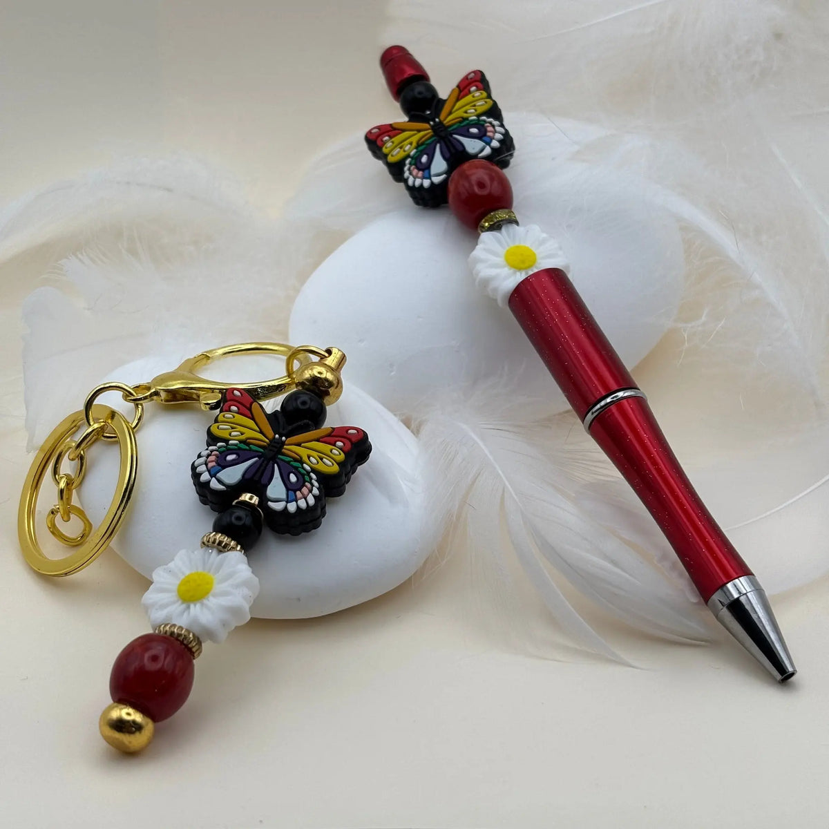 Stylo rouge décoré d’un papillon coloré et fleur blanche, posé à côté d’un porte-clés assorti avec perles rouges et blanches