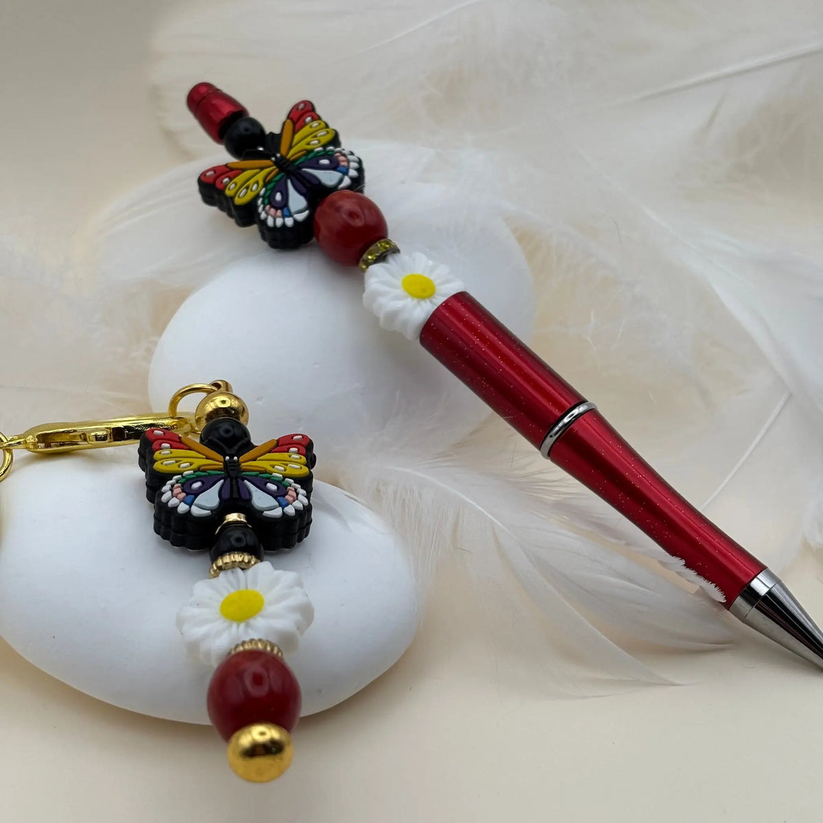 Stylo rouge orné d’un papillon multicolore et perles rouges et blanches, présenté avec un porte-clés assorti décoré d’une fleur blanche