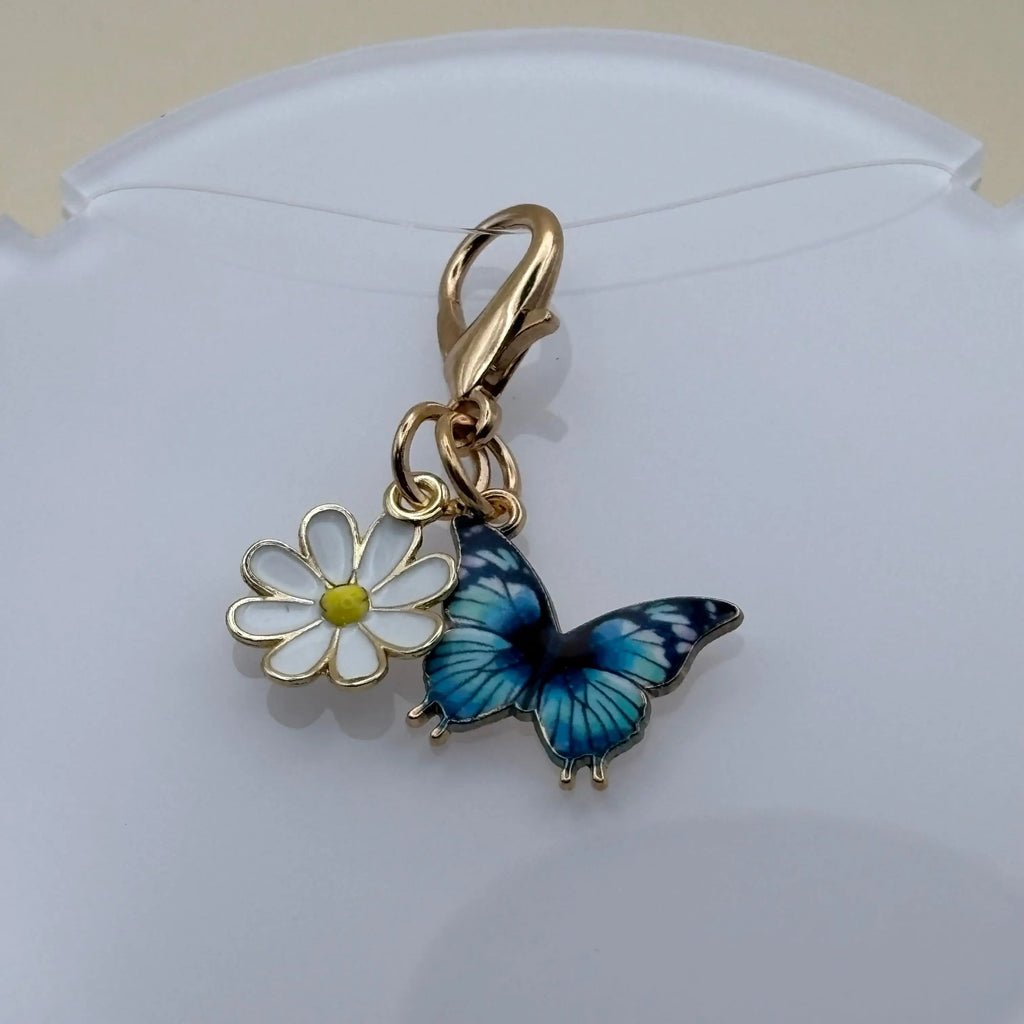 Bijou en acier à accrocher représentant un papillon bleu posé sur une marguerite, évoquant la légèreté, la nature et l’élégance florale.
