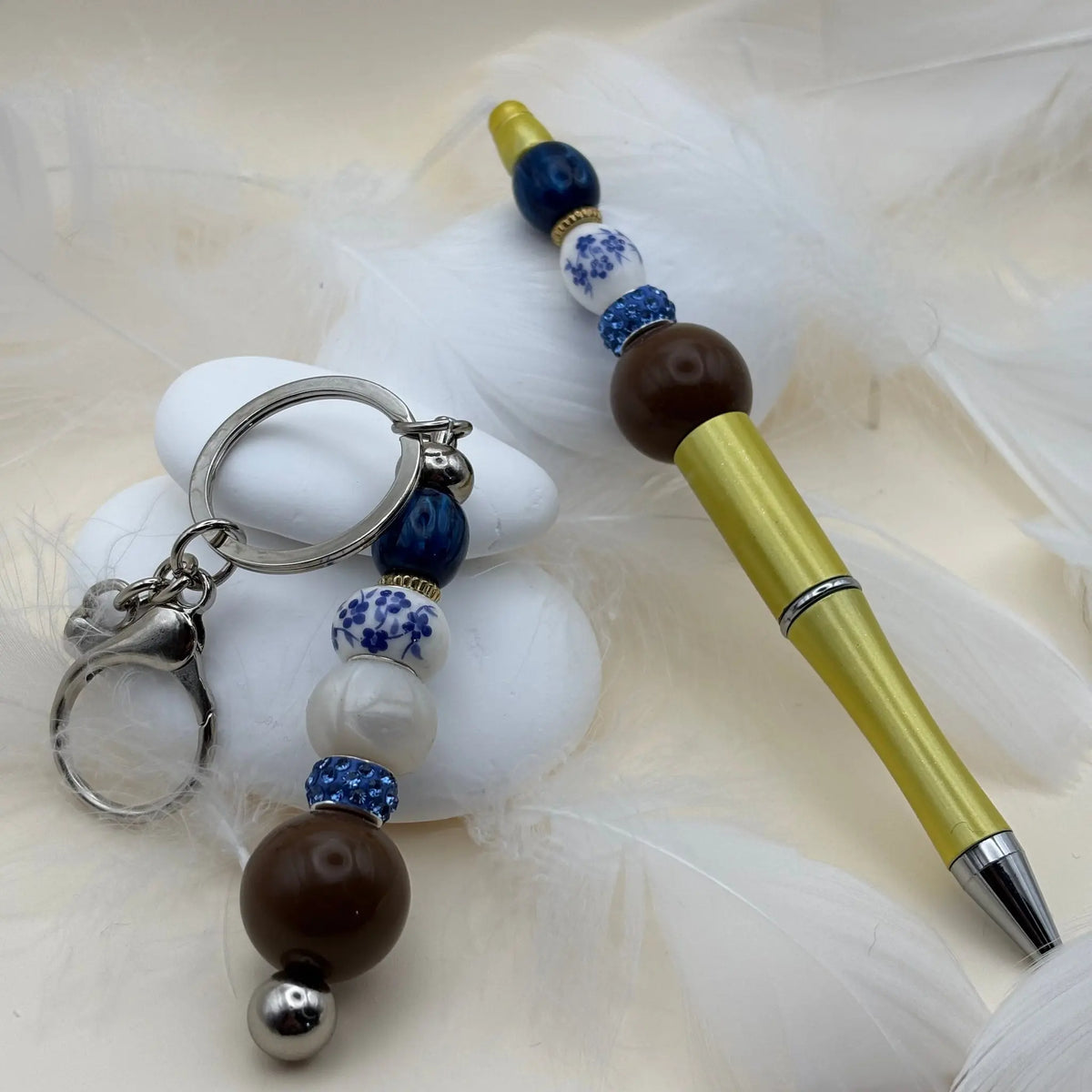 Stylo jaune orné de perles bleues, blanches et marron, style porcelaine, accompagné d’un porte-clés assorti posé sur des galets blancs