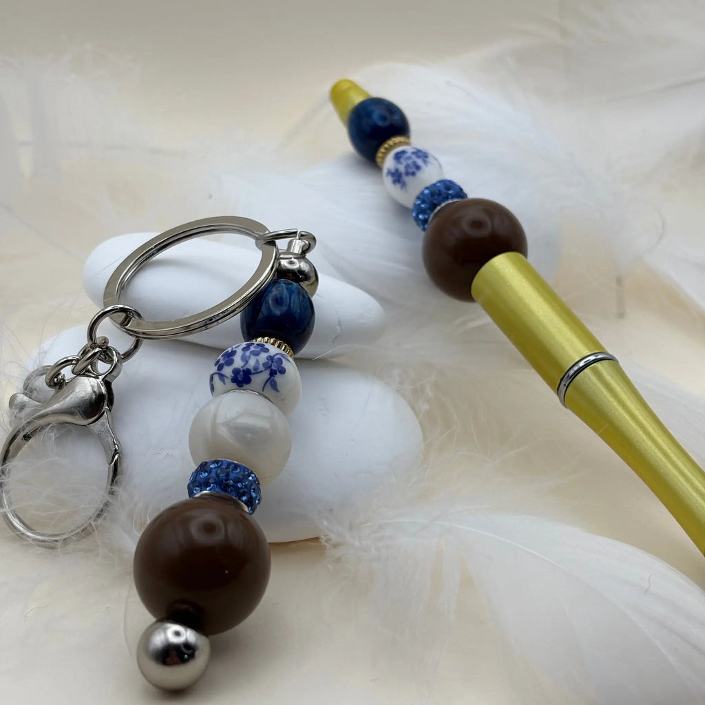 Stylo jaune décoré de perles bleu porcelaine, blanches et marron, accompagné d’un porte-clés assorti sur fond de plumes blanches