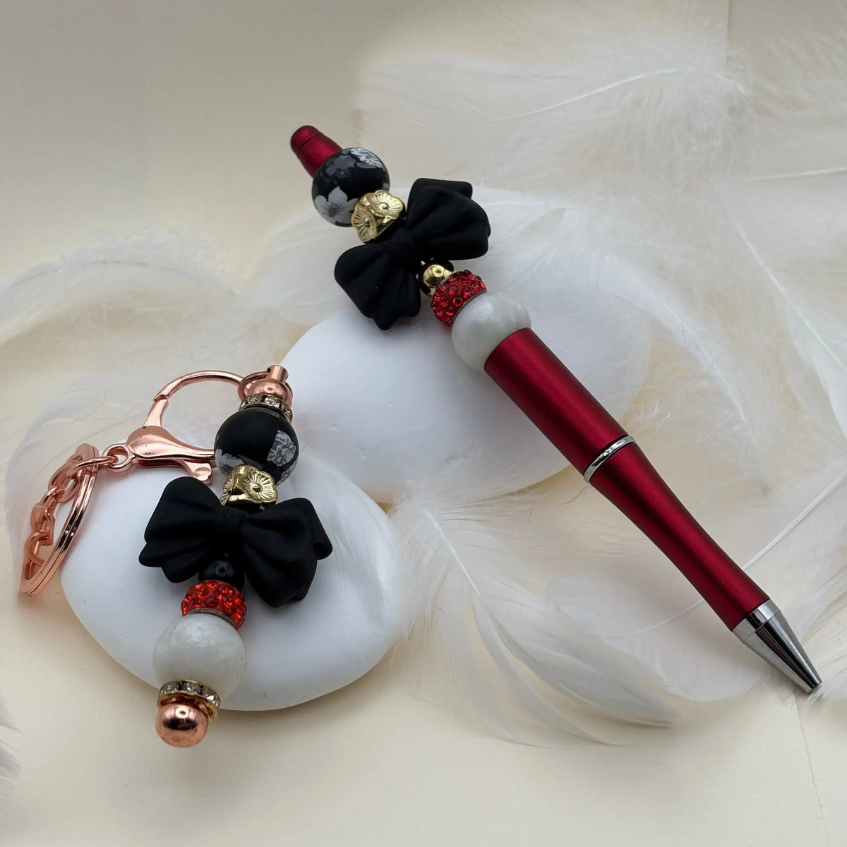 Stylo doré décoré de perles marron et beige avec fleurs crème, posé à côté d’un porte-clés assorti sur des plumes blanches