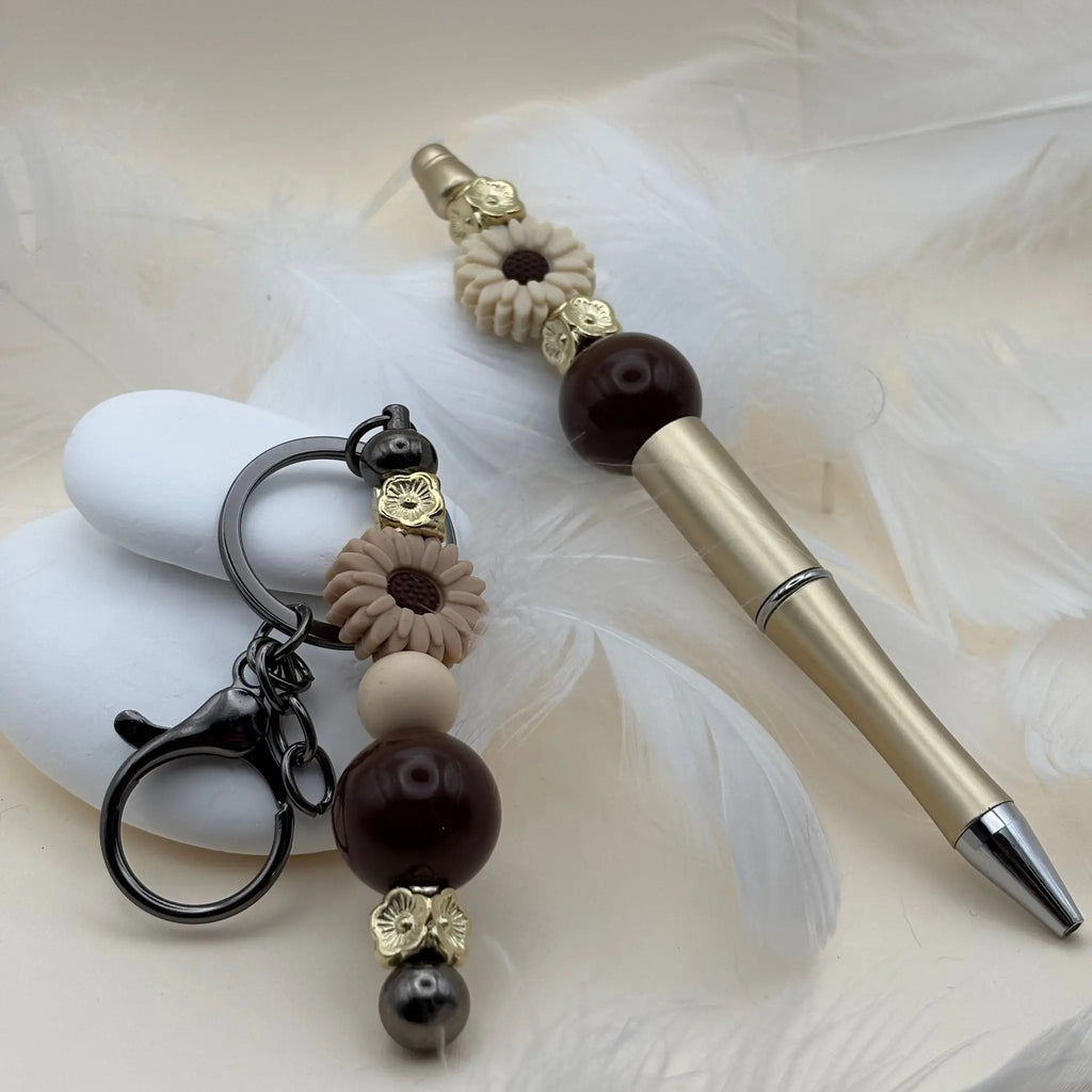 Stylo doré décoré de perles marron et beige avec fleurs en résine crème, accompagné d’un porte-clés assorti posé sur des plumes blanches