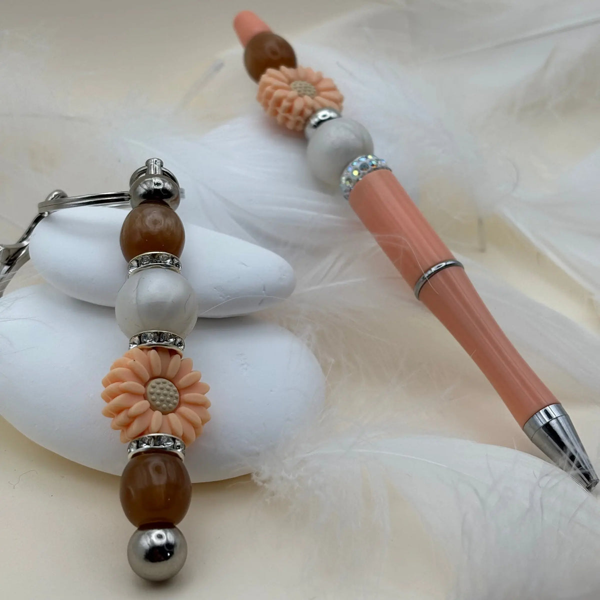 Stylo rose saumon orné de perles beige et marron avec fleurs en résine pêche, présenté avec un porte-clés coordonné sur des galets blancs