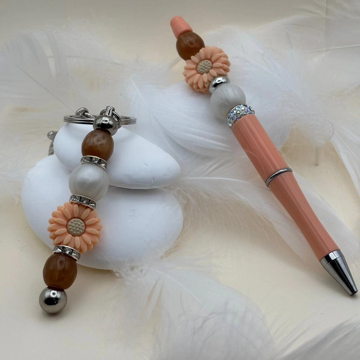 Stylo rose saumon décoré de fleurs pêche et perles blanches et marron, avec un porte-clés assorti sur fond de plumes blanches